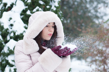 Portrait of young beautiful emotional woman in hooded down coat and gloves on snow covered garden background. Winter snowy landscape