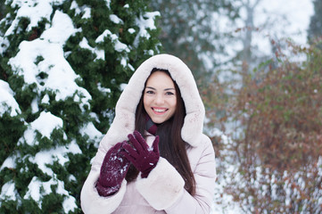 Portrait of young beautiful emotional woman in hooded down coat and gloves on snow covered garden background. Winter snowy landscape