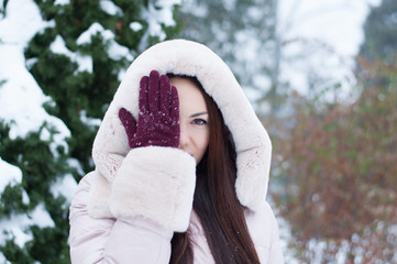 Portrait of young beautiful emotional woman in hooded down coat and gloves on snow covered garden background. Winter snowy landscape