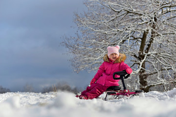 A little girl in a pink down jacket sitting on a sled under a branched tree in the snowy winter.