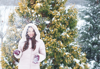 Portrait of young beautiful emotional woman in hooded down coat and gloves on snow covered garden background. Winter snowy landscape