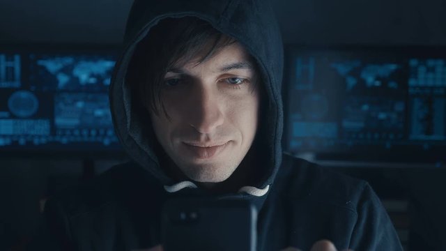 Dangerous Hacker in the hood uses a smartphone in a secret data center filled with screens