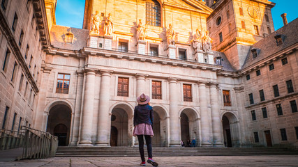 A young woman, tourist in a hat is standing in front of the basilica at El Escorial palace and monastery at the San Lorenzo de El Escorial during sunset. Famous kings residence near Madrid in Spain