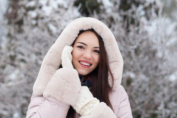 Portrait of young beautiful emotional woman in hooded down coat and gloves on snow covered garden background. Winter snowy landscape