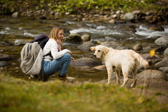 Beautiful blonde teenager girl with backpack in hiking bots and warm clothes, plays with her golden retriever friend outdoor, next to a river.