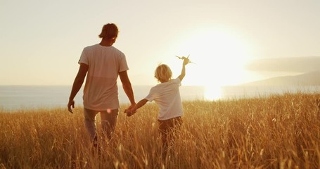 Adorable father and son walking together through golden field at sunset