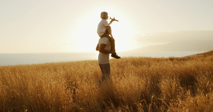 Handsome father spinning adorable son on his shoulders with toy airplane, sunset golden field