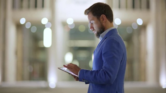 Side view tracking shot of serious businessman using tablet computer while walking down illuminated night street