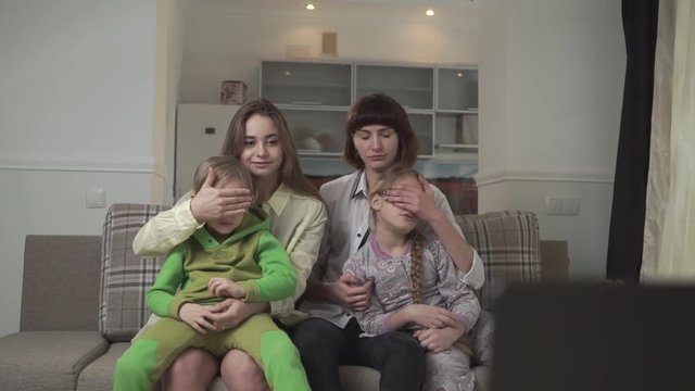 Family watching TV sitting on the couch in the guest room. Older sisters close their eyes to children because of bad inappropriate content on the screen. Native people spend time together. Family