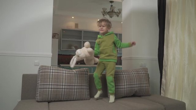 Jouful little boy in green pajamas jumping on the sofa at home. Child grabs pillow and throws in on the floor. Happy cheerful kid. Slow motion.