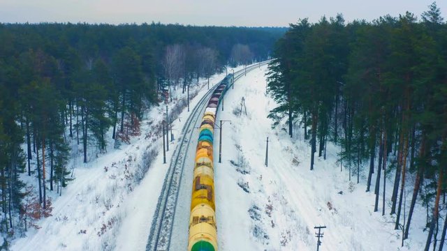 Aerial following view of the freight train, carrying tank cars with oil and cargo containers, moving on the winter snowy forest