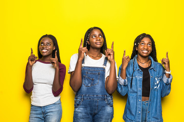 Three beautiful smiling african women pointed up with hands looking up isolated over yellow background