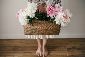 Boho girl holding pink and white peonies in rustic basket and standing barefoot on wooden floor....