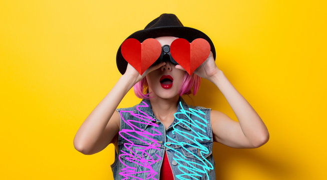 Portrait of young style hipster girl with pink hair style with binoculars with heart shape on yellow background