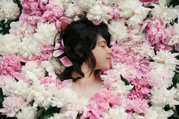 Beautiful brunette girl in many pink and white peonies. Happy boho woman portrait with peony flowers, top view. Creative floral photo. Aroma scent concept. International Womens Day