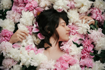 Beautiful brunette girl lying in many pink and white peonies. Boho woman portrait with natural makeup in peony flowers. Creative floral photo. Aroma scent concept. International Womens Day