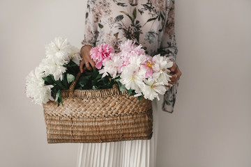 Boho girl holding pink and white peonies in rustic basket. Stylish hipster woman in bohemian floral...