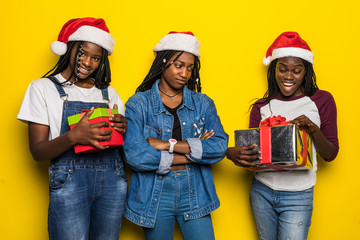 Three young african woman in santa hat share Christmas present on yellow background