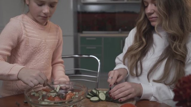 Younger sister helps the older sister coocking vegetable salad for breakfast sitting at a small table in the cozy kitchen. Sisters relationship.