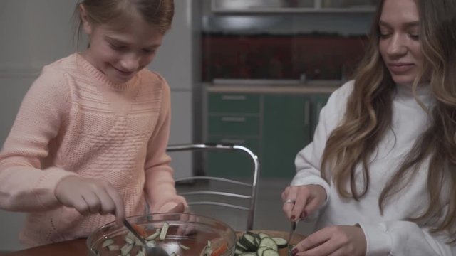 Younger sister helps the older sister coocking vegetable salad for breakfast. Eldest girl slices cucumbers and younger girl one mixes the salad in a plate using a spoon. Sisters relationship.