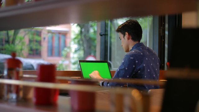 Latin American hispanic young man sitting in coffee shop with a laptop drinking coffee, chroma key screen, depth shot through objects. High quality ProRes 422 HQ