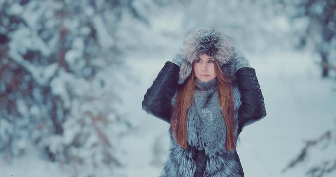 Beautiful girl in a fur hat and jacket walks in the park in winter. Snowfall.