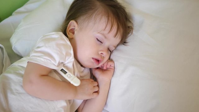 Small child sleeps in hospital ward on white bedding and measures temperature with thermometer. Treatment of children in hospital. Sick baby improves his health in hospital.