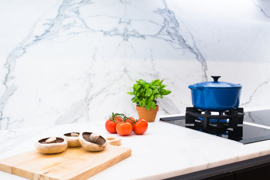 Blue enamel pot cooking shown with ingredients of mushrooms, tomatoes and basil on marble kitchen countertop with marble splash back showing stone slab book match detail