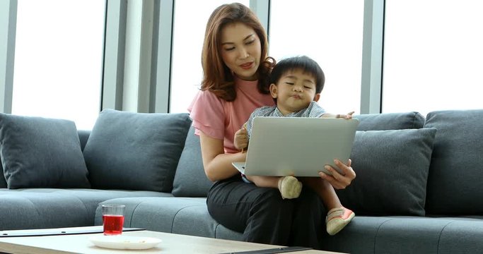 Asian mother using notebook with her son, he sitting on his mother lap, in living room, concept for mother and son spending time together.