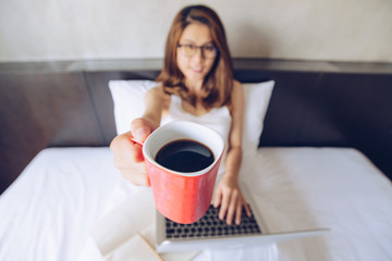 Portrait of Asian woman working on the bed and giving a red cup of coffee to you. Conceptual of woman lifestyle.