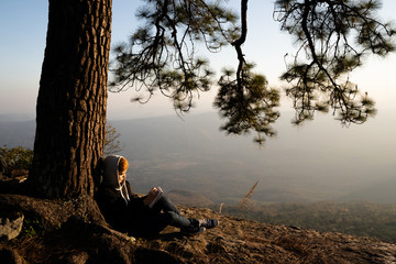 Woman sitting under pine tree reading and writing looking out at beautiful natural view