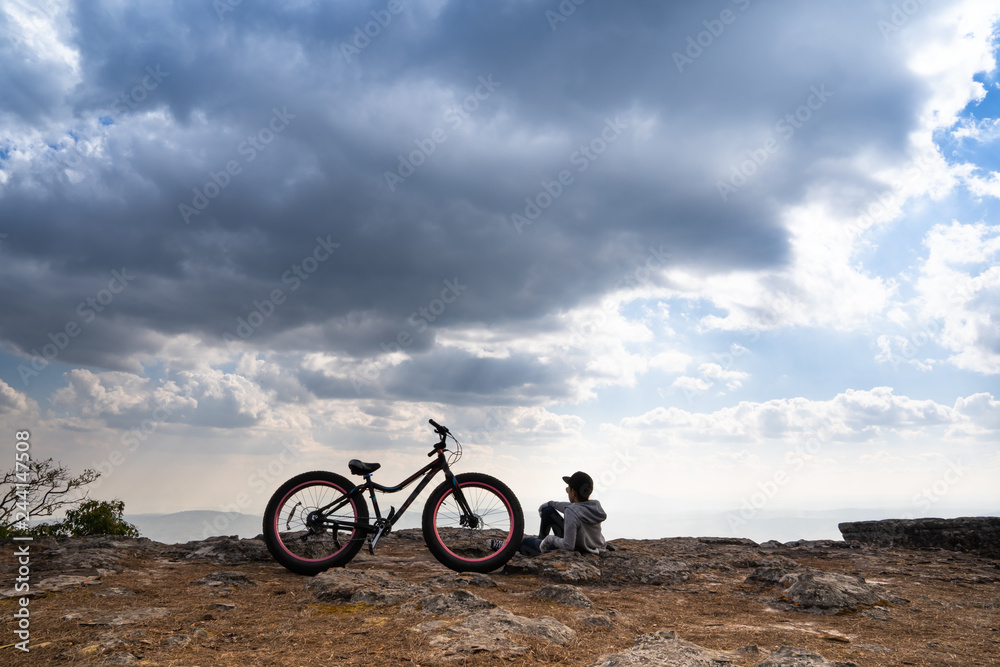 Wall mural A person sitting down beside a bicycle on rocky mountain looking out at scenic natural view and beautiful blue sky - Wall murals