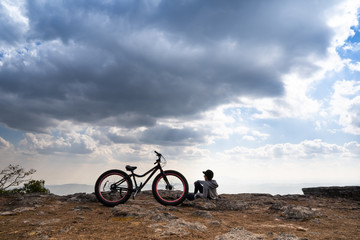 A person sitting down beside a bicycle on rocky mountain looking out at scenic natural view and beautiful blue sky