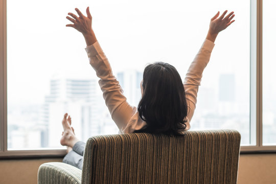 Life quality concept with business woman rear view relaxing sitting in rest on cozy armchair in modern hotel guest room or luxury home living room looking toward beautiful city urban scene