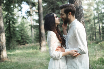 Naklejka premium Young beautiful Slavic couple in love walks through the woods, sweet girl in a simple boho-style wedding dress. They are happy and calm. Golden hour, summer day, warm. They kiss, hug and hold hands.
