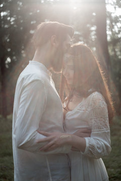 Young beautiful Slavic couple in love walks through the woods, sweet girl in a simple boho-style wedding dress. They are happy and calm. Golden hour, summer day, warm. They kiss, hug and hold hands.