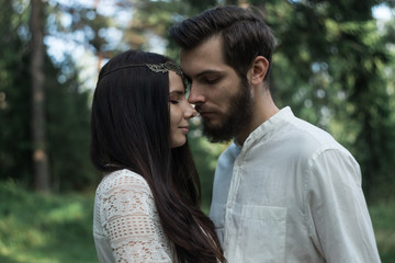 Young beautiful Slavic couple in love walks through the woods, sweet girl in a simple boho-style wedding dress. They are happy and calm. Golden hour, summer day, warm. They kiss, hug and hold hands.