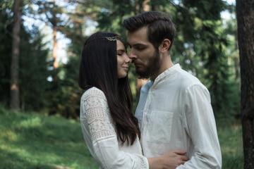 Fototapeta premium Young beautiful Slavic couple in love walks through the woods, sweet girl in a simple boho-style wedding dress. They are happy and calm. Golden hour, summer day, warm. They kiss, hug and hold hands.
