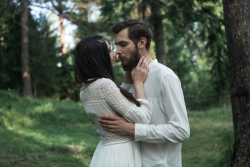 Young beautiful Slavic couple in love walks through the woods, sweet girl in a simple boho-style wedding dress. They are happy and calm. Golden hour, summer day, warm. They kiss, hug and hold hands.