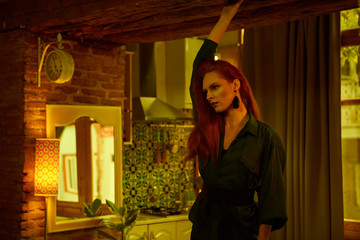Ginger lady posing in loft apartment in Tbilisii. She wears green shirt and black jeans. Her hair is curly. Flat has big window. Walls are of red bricks. Photo has vintage style and is yellowish.