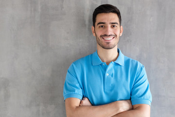 Portrait of attractive young man in blue polo shirt, standing with crossed arms against gray...