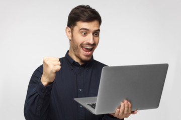 Excited young businessman in blue shirt looking at laptop screen isolated on gray background