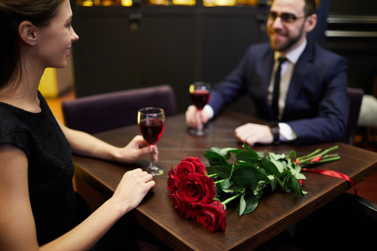 Young amorous couple with glasses of red wine sitting by table in luxurious restaurant opposite each other