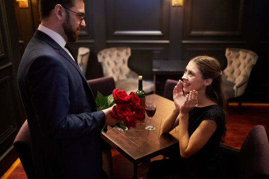 Cheerful girl looking at young elegant man in suit with bunch of red roses standing in front