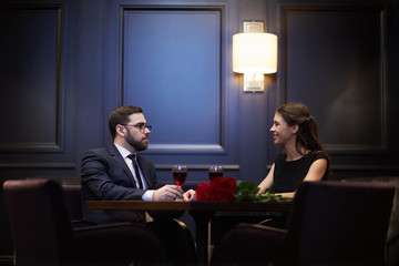Young romantic dates sitting by table opposite one another while enjoying talk by dinner in restaurant