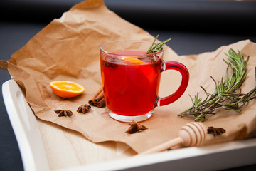 Transparent glass mug with fragrant black tea, with anise, rosemary and orange on a tray on a dark background. Next to the mug is kraft paper, dried anise flowers, cinnamon and honey dipper
