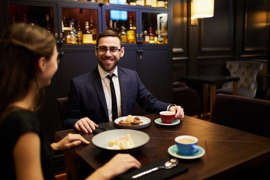 Cheerful young man talking to his girlfriend in luxurious restaurant while both having dessert