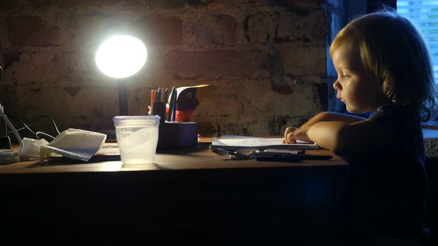 Kid lefty is drawing in the album. Little girl, child sits near the window and painting