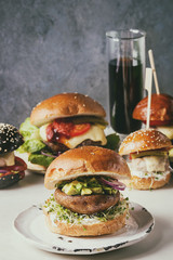 Variety of homemade classic, vegan and mini burgers in wheat and black buns with beef and veal cutlets, portobello mushroom, avocado on white ceramic board over white marble table. With glass of cola.