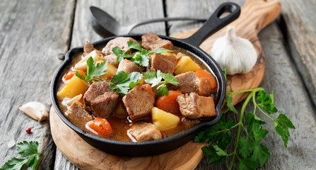 Meat stewed with potatoes, carrots and spices in iron pan on wooden background . - obrazy, fototapety, plakaty